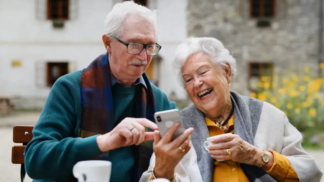 Uśmiechnięta para seniorów korzysta z telefonu, odkrywając nowe aplikacje dla seniorów. Radość i wspólne chwile przy kawie.