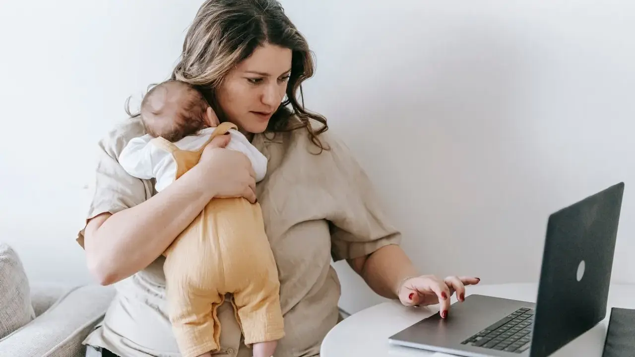Zmęczona kobieta z dzieckiem na rękach pracuje przy laptopie. To obraz zespołu stresu opiekuna, gdzie obowiązki domowe i zawodowe nakładają się na siebie.