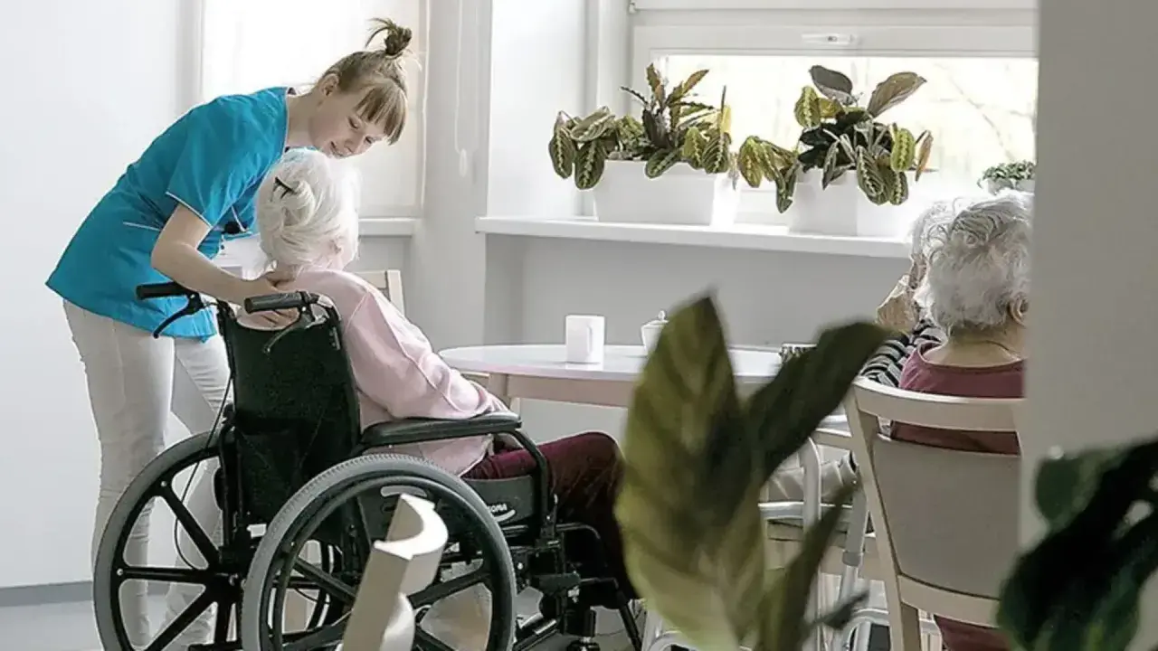 Opiekunka pomaga pacjentowi geriatrycznemu w wózku inwalidzkim. W tle siedzą inne starsze osoby.