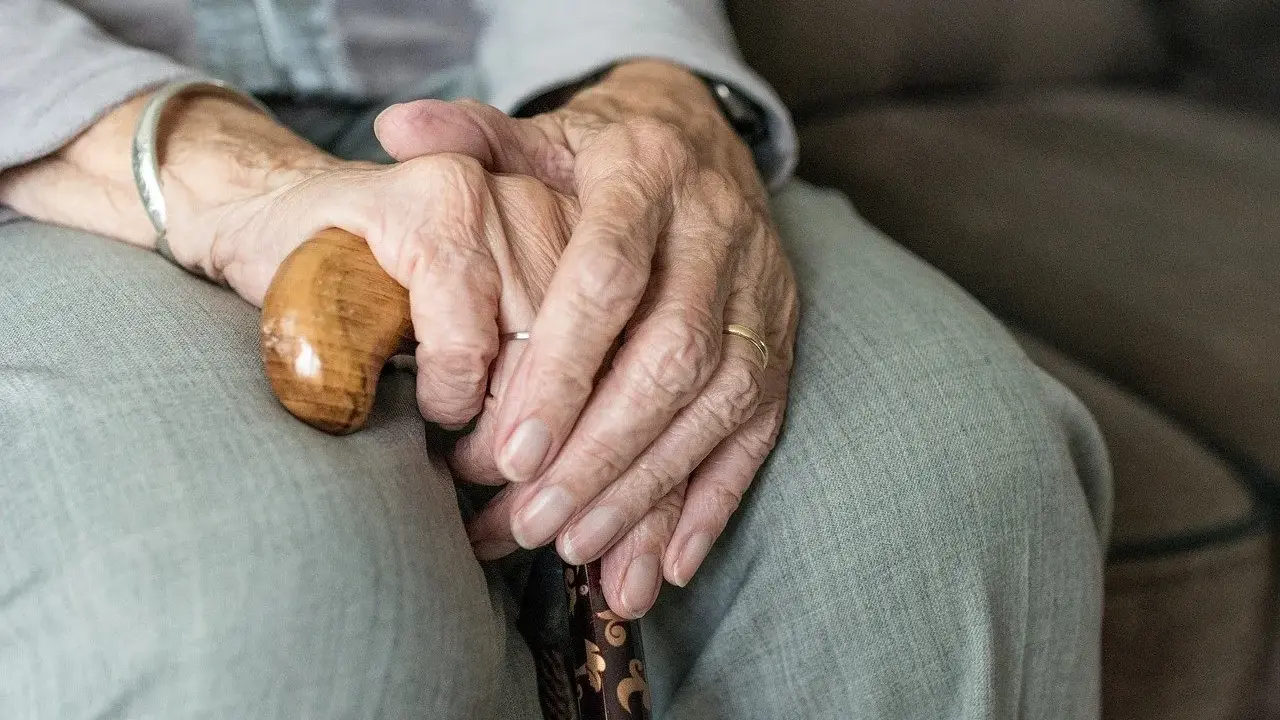 Dłonie starszej osoby spoczywają na lasce. Zmarszczki i obrączki symbolizują długie życie, a trudności z oddawaniem moczu mogą być jego częścią.