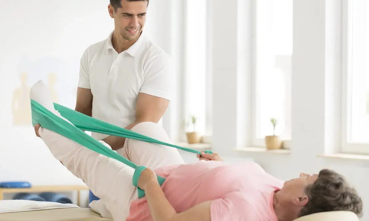 Fizjoterapeuta pomaga pacjentce w rehabilitacji poszpitalnej po endoprotezie biodra NFZ, ćwicząc nogę z taśmą oporową.
