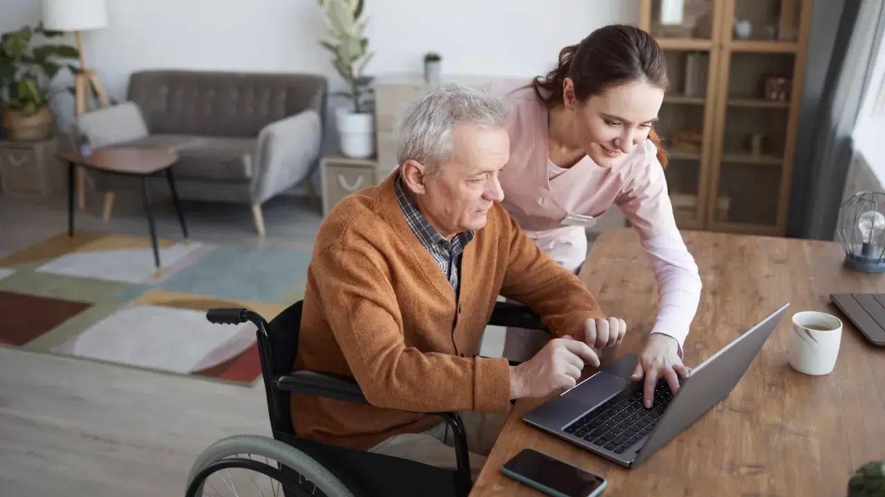 Starszy pan na wózku inwalidzkim z pomocą opiekunki korzysta z laptopa. Nowoczesna technologia wspomagająca ułatwia mu codzienne życie.