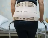 Osoba w czarnych legginsach i czarnym topie, wspierająca się o chodzik. Widać wyraźnie, że gorset ortopedyczny nie jest widoczny spod ubrania.