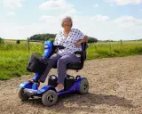 Uśmiechnięta kobieta na niebieskim skuterze elektrycznym inwalidzkim, ciesząca się dniem na wsi.