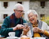 Uśmiechnięta para seniorów korzysta z telefonu, odkrywając nowe aplikacje dla seniorów. Radość i wspólne chwile przy kawie.