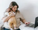 Zmęczona kobieta z dzieckiem na rękach pracuje przy laptopie. To obraz zespołu stresu opiekuna, gdzie obowiązki domowe i zawodowe nakładają się na siebie.
