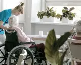 Opiekunka pomaga pacjentowi geriatrycznemu w wózku inwalidzkim. W tle siedzą inne starsze osoby.