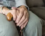Dłonie starszej osoby spoczywają na lasce. Zmarszczki i obrączki symbolizują długie życie, a trudności z oddawaniem moczu mogą być jego częścią.