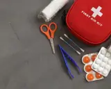 Czerwona apteczka pierwszej pomocy z krzyżem. Obok nożyczki, pęseta, waciki, bandaż i tabletki. Pamiętaj, czego nie powinna zawierać apteczka pierwszej pomocy.