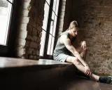 Mężczyzna w sportowym stroju siedzi przy oknie, masując nogę. Czas zrostu więzozrostu, być może po kontuzji.