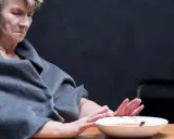 Starsza osoba w szarym szalu odwraca się od talerza z zupą, wyraźnie pokazując, że osoba leżąca nie chce jeść.