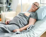 Starsza kobieta w okularach leży w łóżku szpitalnym, otulona kocem. Pokój wyposażony jest w udogodnienia dla osób leżących, w tle widać kule.