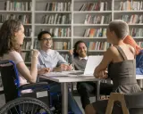 Grupa studentów, w tym osoba na wózku inwalidzkim (3 stopień niepełnosprawności), dyskutuje przy stole w bibliotece.