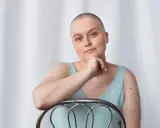 Kobieta po mastektomii, z łysą głową, opiera się o krzesło. Myśli o tym, jak uzyskać biustonosz po mastektomii refundacja.