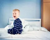 Mały chłopiec w piżamie siedzi na łóżku i płacze. Zastanawiasz się, dlaczego dziecko popuszcza w majtki?