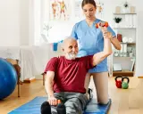 Starszy pan ćwiczy z fizjoterapeutką, podnosząc hantle. To przykład, dla kogo przeznaczony jest bon senioralny – na rehabilitację i aktywność.