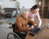 Starszy pan na wózku inwalidzkim z pomocą opiekunki korzysta z laptopa. Nowoczesna technologia wspomagająca ułatwia mu codzienne życie.