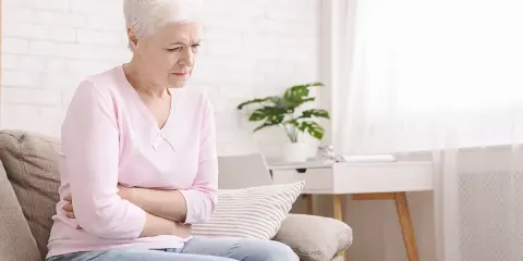 Starsza kobieta siedzi na kanapie, trzymając się za brzuch. Szuka rozwiązania co na zaparcia u starszej osoby.