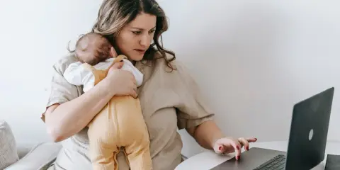 Zmęczona kobieta z dzieckiem na rękach pracuje przy laptopie. To obraz zespołu stresu opiekuna, gdzie obowiązki domowe i zawodowe nakładają się na siebie.