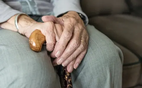 Dłonie starszej osoby spoczywają na lasce. Zmarszczki i obrączki symbolizują długie życie, a trudności z oddawaniem moczu mogą być jego częścią.