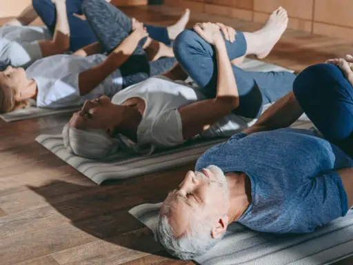 Grupa seniorów 60 plus ćwiczy na macie. Wszyscy leżą na plecach, przyciągając jedno kolano do klatki piersiowej.