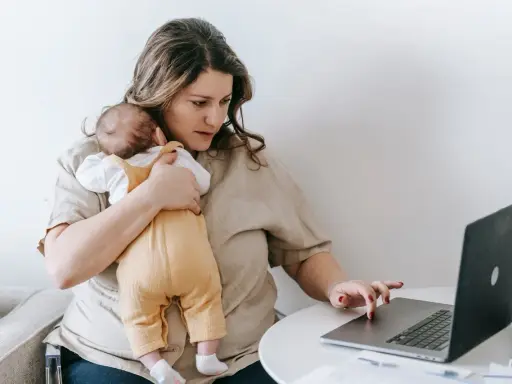 Zmęczona kobieta z dzieckiem na rękach pracuje przy laptopie. To obraz zespołu stresu opiekuna, gdzie obowiązki domowe i zawodowe nakładają się na siebie.