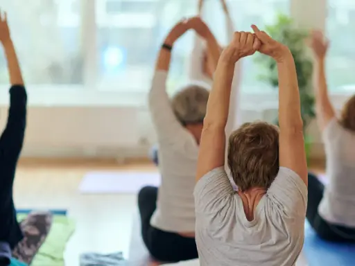Grupa seniorów ćwiczy jogę, podnosząc ręce do góry. Wszyscy siedzą na matach, skupieni na ruchu.