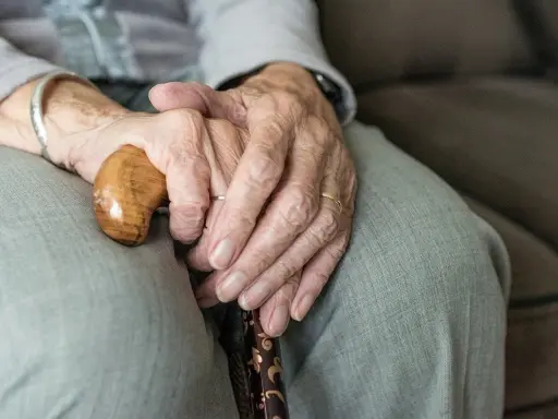 Dłonie starszej osoby spoczywają na lasce. Zmarszczki i obrączki symbolizują długie życie, a trudności z oddawaniem moczu mogą być jego częścią.