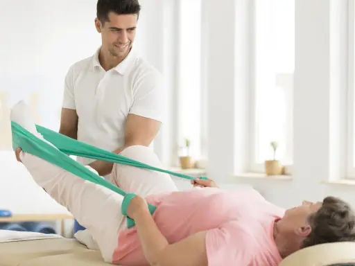 Fizjoterapeuta pomaga pacjentce w rehabilitacji poszpitalnej po endoprotezie biodra NFZ, ćwicząc nogę z taśmą oporową.
