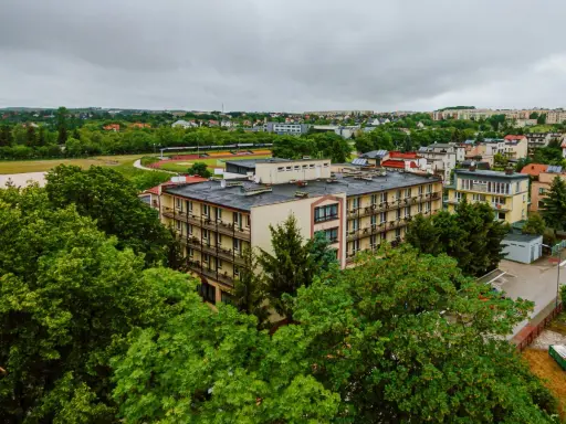 Budynek rehabilitacyjny otoczony zielenią, z widokiem na miasto i stadion.