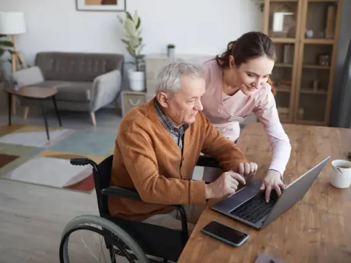 Starszy pan na wózku inwalidzkim z pomocą opiekunki korzysta z laptopa. Nowoczesna technologia wspomagająca ułatwia mu codzienne życie.