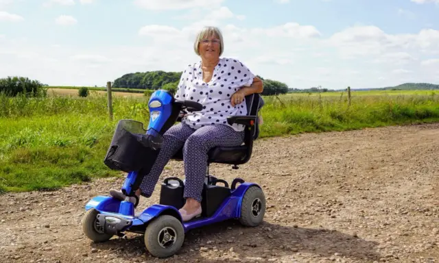 Uśmiechnięta kobieta na niebieskim skuterze elektrycznym inwalidzkim, ciesząca się dniem na wsi.