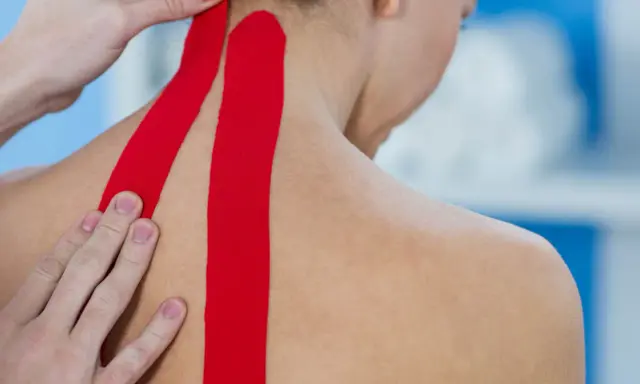 Czerwone plastry kinesio taping na karku i ramieniu, aplikowane przez fizjoterapeutę.