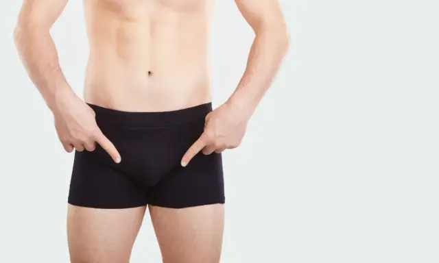 Mężczyzna w czarnych bokserkach wskazuje palcami na okolice krocza, sugerując ćwiczenia na mięśnie kegla u mężczyzn.