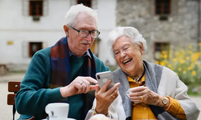 Uśmiechnięta para seniorów korzysta z telefonu, odkrywając nowe aplikacje dla seniorów. Radość i wspólne chwile przy kawie.