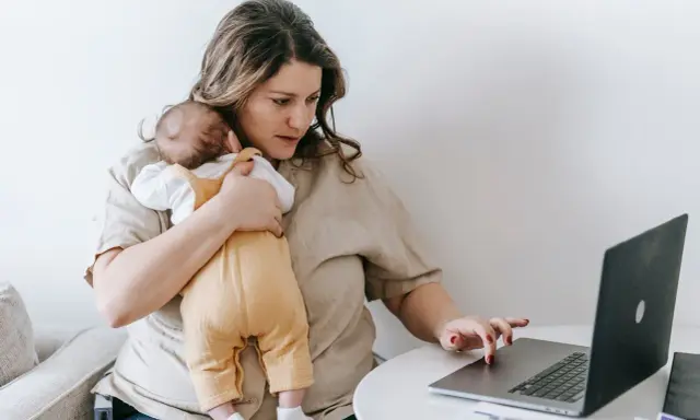 Zmęczona kobieta z dzieckiem na rękach pracuje przy laptopie. To obraz zespołu stresu opiekuna, gdzie obowiązki domowe i zawodowe nakładają się na siebie.