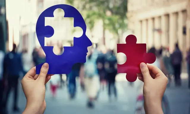 Dwie ręce trzymają elementy układanki: niebieski profil głowy z brakującym fragmentem i czerwony element. Symbolizuje to etapy demencji starczej.
