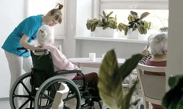 Opiekunka pomaga pacjentowi geriatrycznemu w wózku inwalidzkim. W tle siedzą inne starsze osoby.