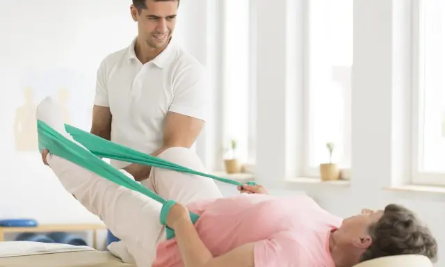 Fizjoterapeuta pomaga pacjentce w rehabilitacji poszpitalnej po endoprotezie biodra NFZ, ćwicząc nogę z taśmą oporową.
