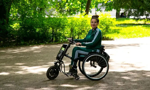 Kobieta w wózku inwalidzkim z napędem elektrycznym, który mógł zostać sfinansowany dzięki dofinansowaniu do wózka inwalidzkiego, cieszy się spacerem w parku.