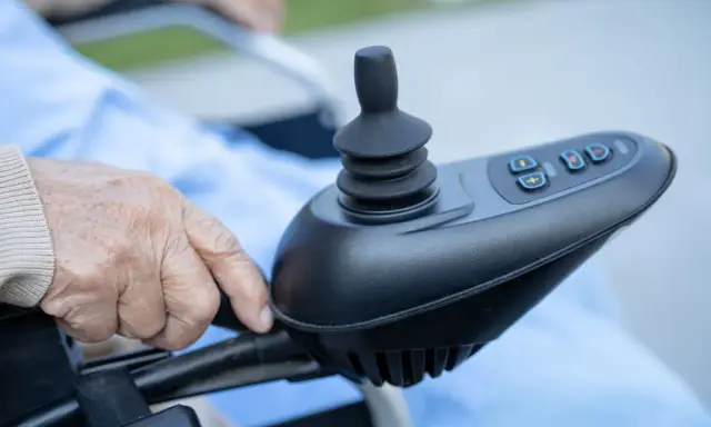 Dłoń starszej osoby na joysticku wózka inwalidzkiego. Czy rozważasz vitafon refundacja NFZ?