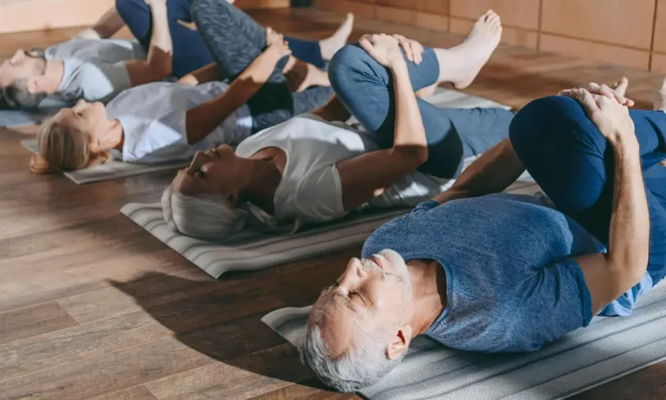 Grupa seniorów 60 plus ćwiczy na macie. Wszyscy leżą na plecach, przyciągając jedno kolano do klatki piersiowej.