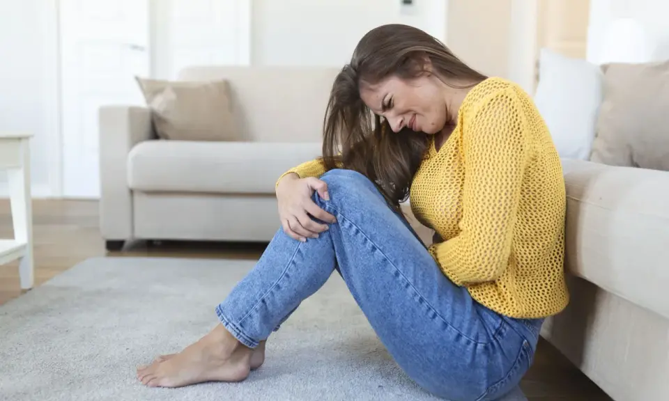 Kobieta siedzi na podłodze, przytulając kolana, z wyrazem bólu na twarzy. Czuje ciągłe parcie na pęcherz, co sprawia jej dyskomfort.