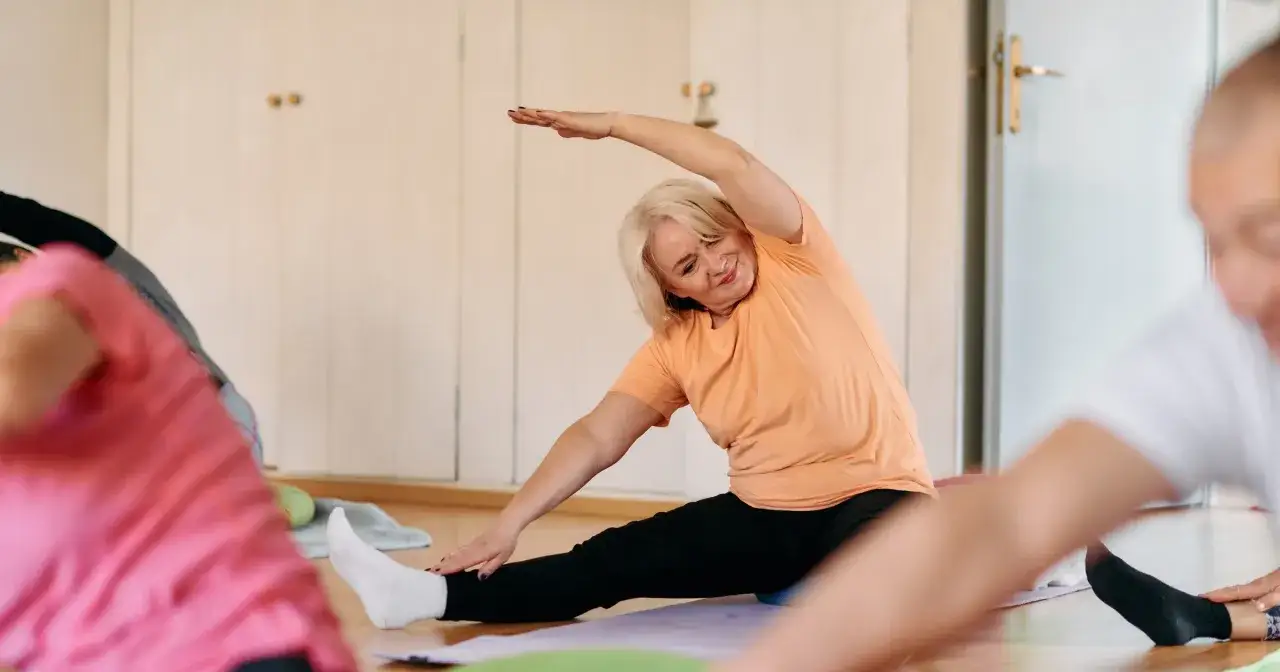 Uśmiechnięta kobieta wykonuje ćwiczenia rozciągające podczas zajęć gimnastyka dla seniorów 60 plus.