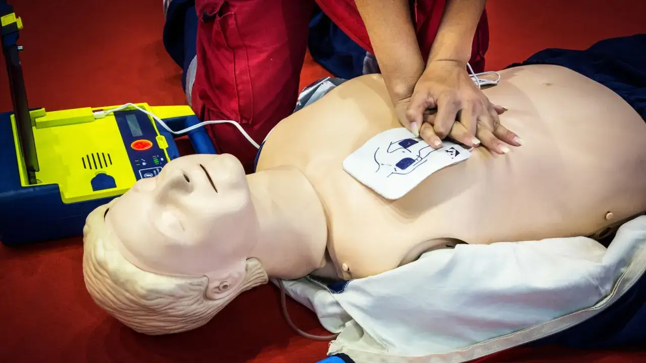 Ćwiczenie RKO z użyciem AED. Manekin do resuscytacji, żółto-niebieski AED, osoba w czerwonych spodniach wykonuje uciśnięcia.