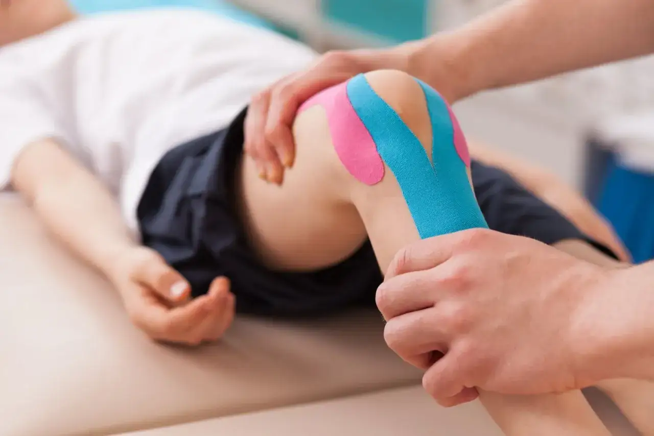 Fizjoterapeuta zakłada kolorowe plastry na kolano dziecka. To kinesio taping, metoda wspierająca rehabilitację.