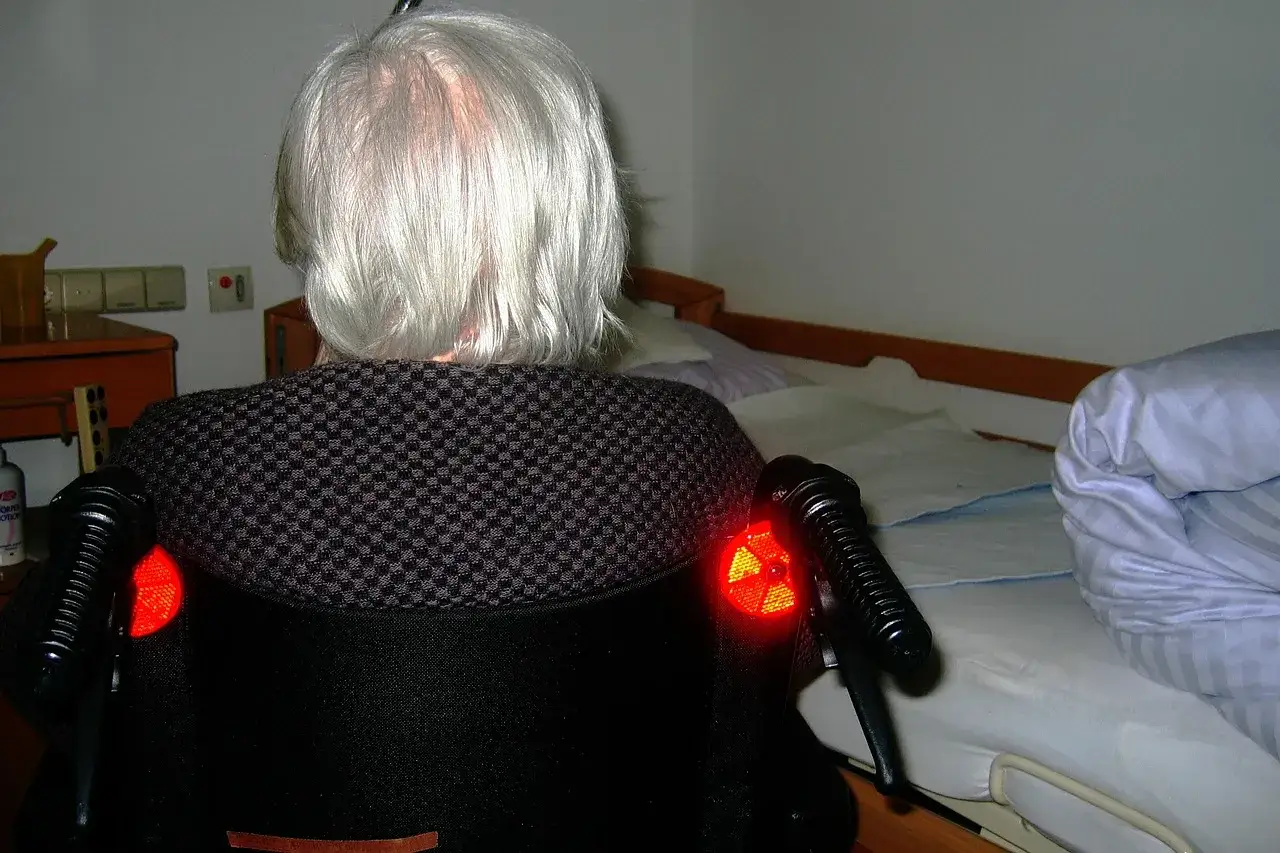 Osoba w fotelu, z siwymi włosami, czeka na opiekę długoterminową. Obok łóżko.