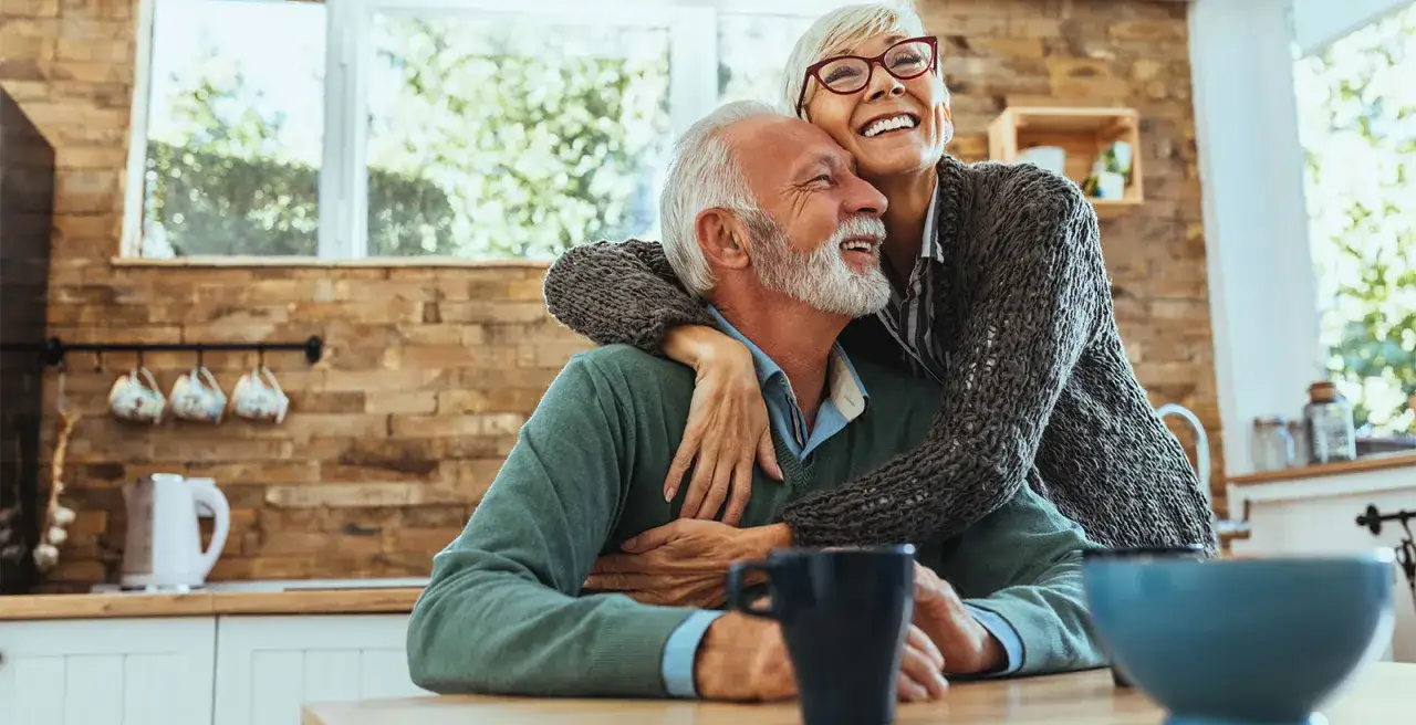 Uśmiechnięta kobieta obejmuje mężczyznę, który mimo że chory na alzheimera, cieszy się chwilą.