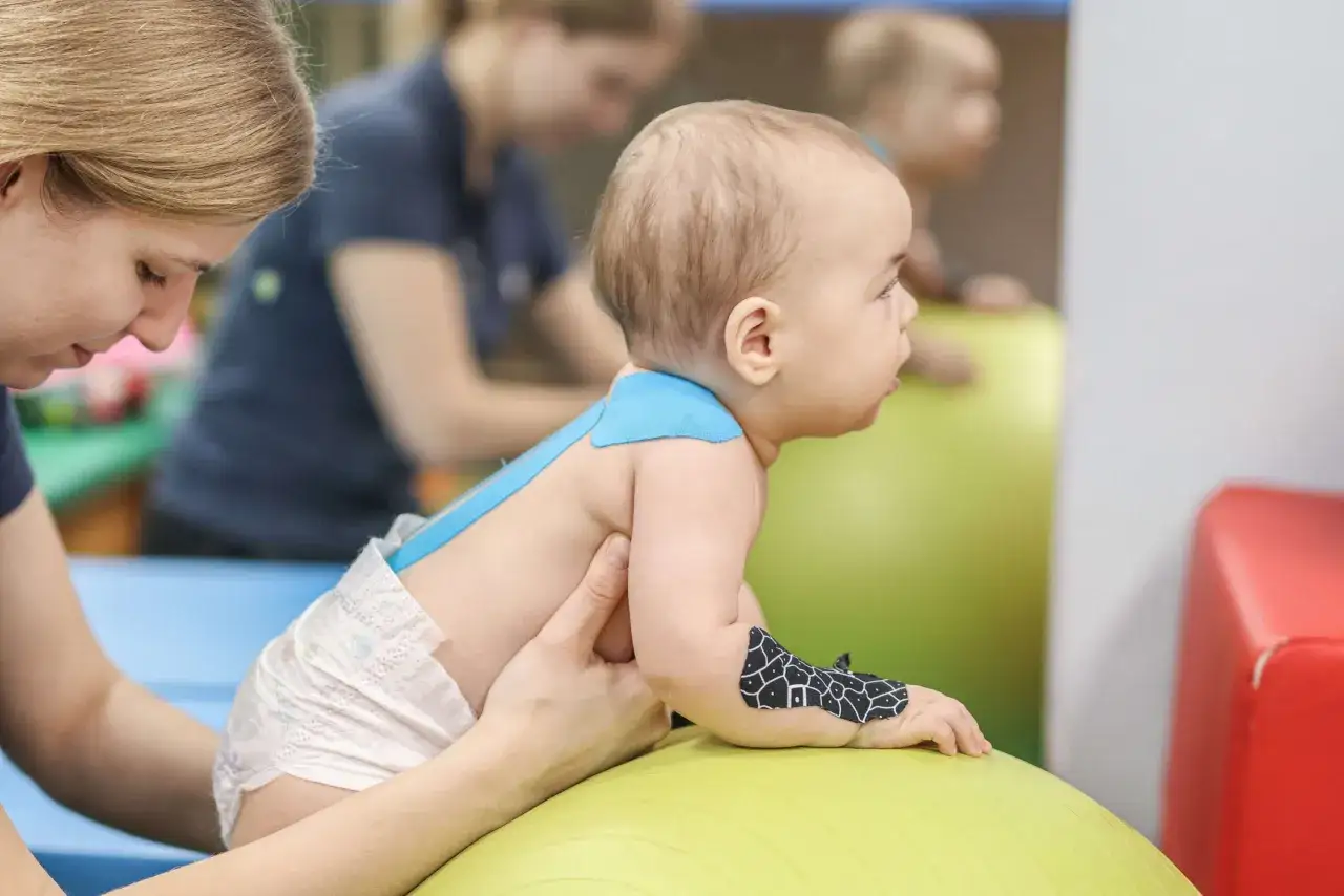 Fizjoterapeuta dla niemowląt delikatnie wspiera dziecko na piłce rehabilitacyjnej. Na plecach malucha widać niebieskie plastry kinesiotaping.